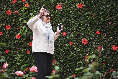 Buy stock photo Senior woman, headphones and dancing in outdoors, smartphone and streaming radio or playlist. Elderly person, flower wall and rock or pop music in retirement, garden and freedom or connection to tech