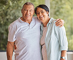 Portrait of a senior happy couple smiling, standing at home in front of a window looking at the camera. Mature hispanic man and woman sharing romantic embrace and showing affection