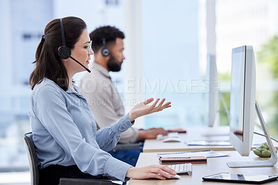 One caucasian call centre telemarketing agent talking on a headset while working on a computer in an office with colleague in the background. Female consultant operating a helpdesk for customer service sales support