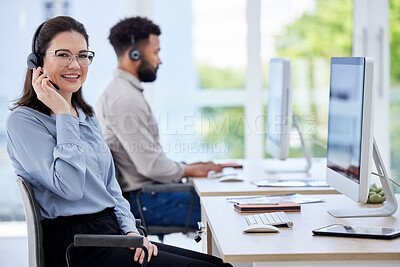 Portrait of one happy caucasian call centre telemarketing agent talking on a headset while working on a computer in an office with colleague in the background. Confident friendly female consultant operating a helpdesk for customer service sales support