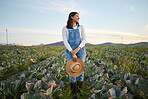 Woman farmer standing in a cabbage field on a farm. Young brunette female with a straw hat and rubber boots looking over a field of organic vegetables