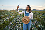 Woman farmer taking a selfie on her smartphone while standing in a cabbage field. Young brunette female with a straw hat using her mobile device on an organic vegetable farm