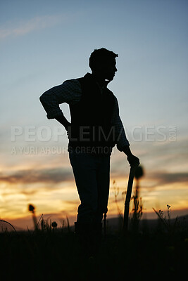 Buy stock photo Silhouette of a male farmer standing in a field with a rake. A man getting ready to work on the farm in the early morning
