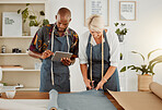 Two designers using a digital tablet while working with material at work. Young african american tailor talking and holding a digital tablet while working with a female caucasian colleague. Mature businesswoman cutting material