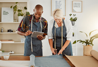 Buy stock photo Two designers using a digital tablet while working with material at work. Young african american tailor talking and holding a digital tablet while working with a female caucasian colleague. Mature businesswoman cutting material