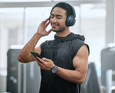 Smiling asian trainer alone in gym, listening to music on headphones from cellphone. Handsome coach standing in health club workout, selecting song from playlist on technology. Man in fitness centre