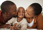 Portrait of adorable little african american boy lying between his mother and father on a bed at home home. Loving parents kissing their toddler son on his cheeks