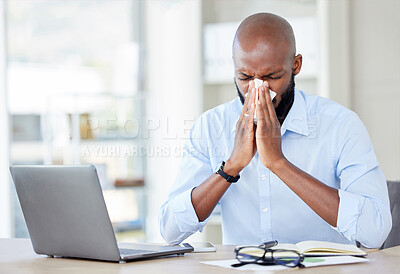 Young african american businessman blowing his nose with a tissue while sitting at a desk at work. Sick male businessperson using a laptop while suffering from allergies at work