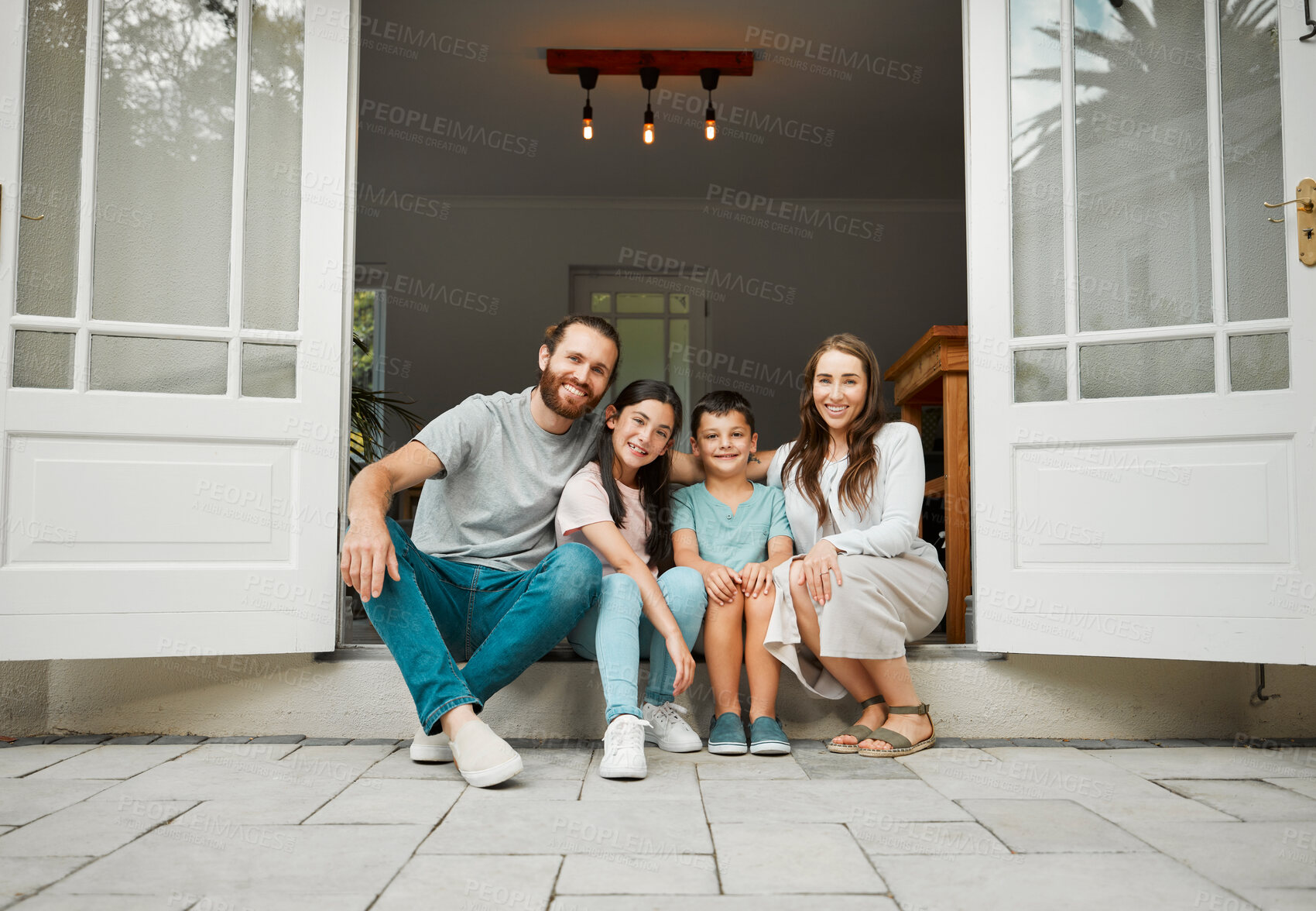 Buy stock photo Portrait of a happy young caucasian family of four sitting at their front door smiling and looking at the camera. Two parents sitting with their little son and daughter in front of their house