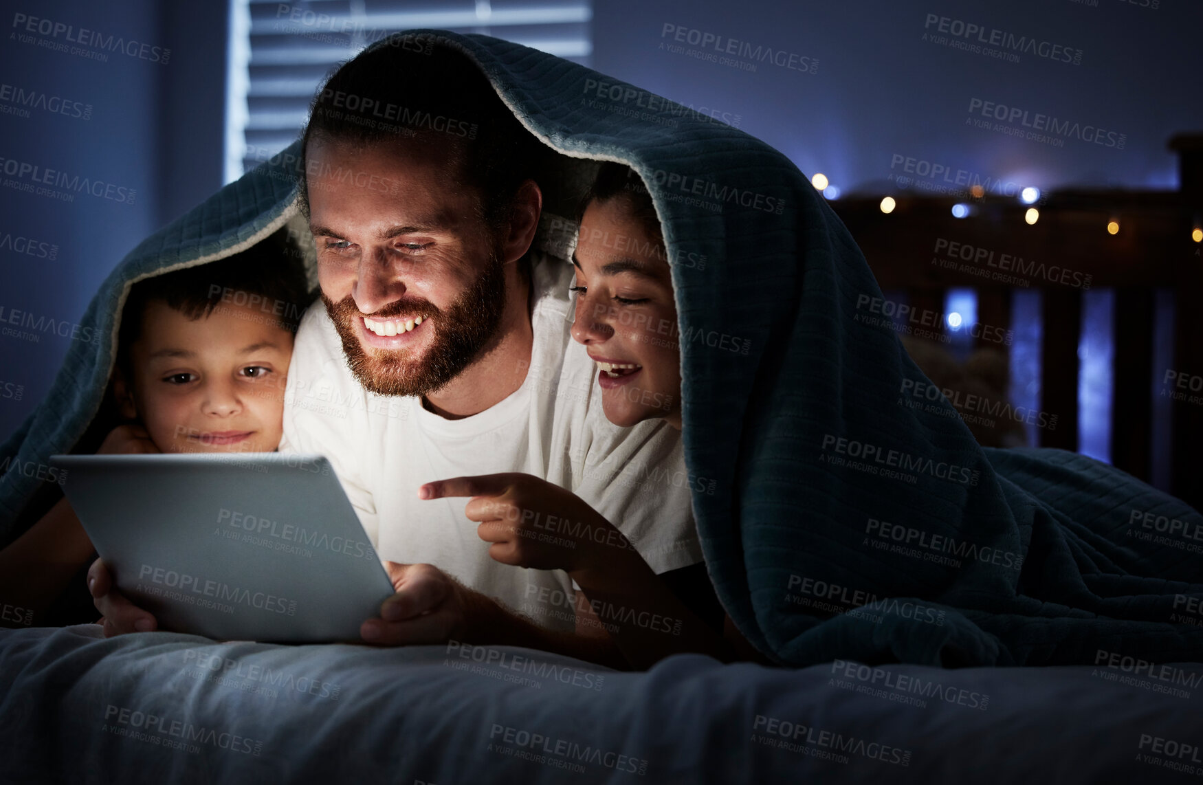 Buy stock photo Happy caucasian family single dad with two children using digital tablet lying under blanket in the dark at night with their faces illuminated by device screen light. Father reading online story or watching video with daughter and son at night