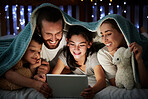 Happy caucasian family with two children using digital tablet lying under blanket in the dark at night with their faces illuminated by device screen light. Family of four reading online story or watching video before bedtime