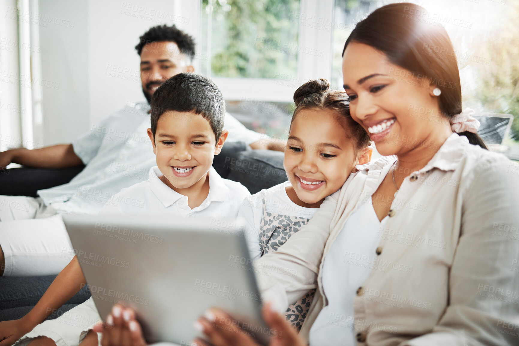 Buy stock photo Happy mixed race smiling family of four bonding on a sofa while streaming movies on the internet using a digital tablet at home. Young couple looking cheerful with their daughter and son