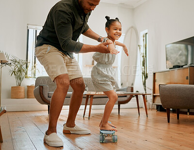 A happy mixed race family of two playing and skating on the lounge floor together. Loving black single parent bonding with his daughter while teaching her how to skate on a skateboard at home