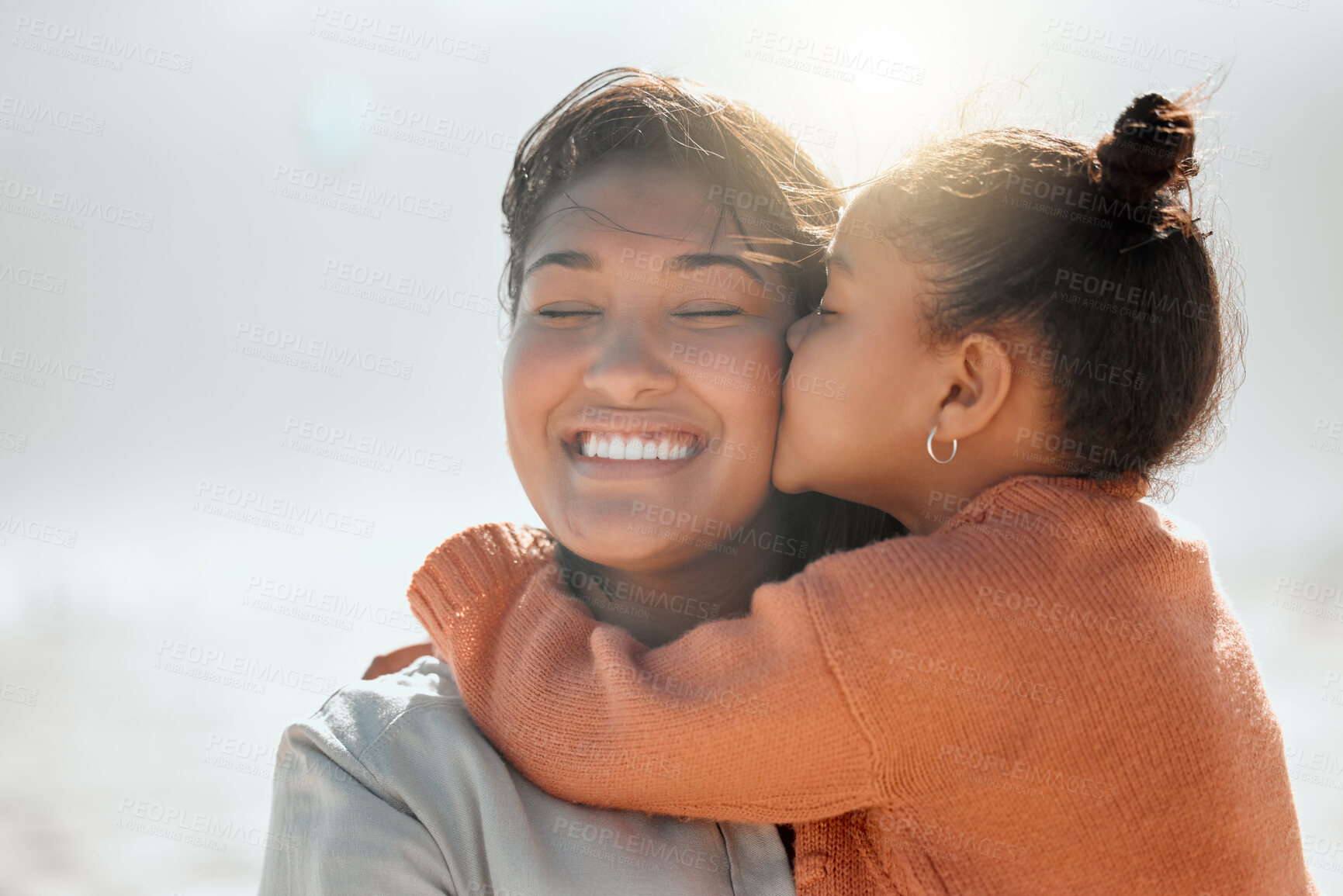 Buy stock photo Adorable mixed race girl kisses her mother on the cheek and outs her arms around her while at the beach. Little daughter showing mommy love and affection while she closes her her eyes and smile