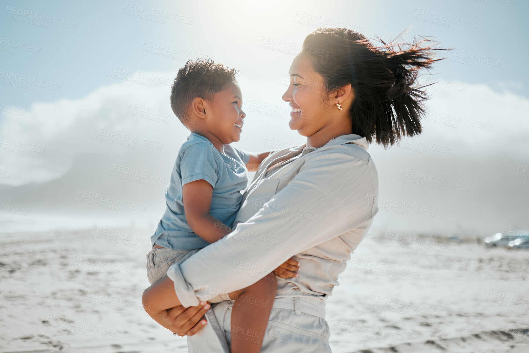 Buy stock photo Loving young mixed race mother holding her adorable little son as they looking into each others eyes on a windy day at the beach. Mother and son spending time and having fun together while on holiday