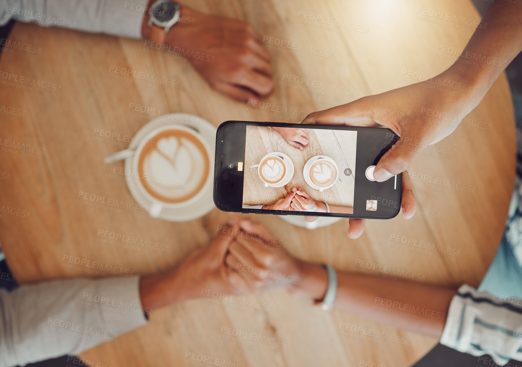 Buy stock photo Close up of device screen while couple holds hands with two cups of coffee on a wooden table while taking a selfie on smartphone. Female and male hands on a table while on a date at a cafe