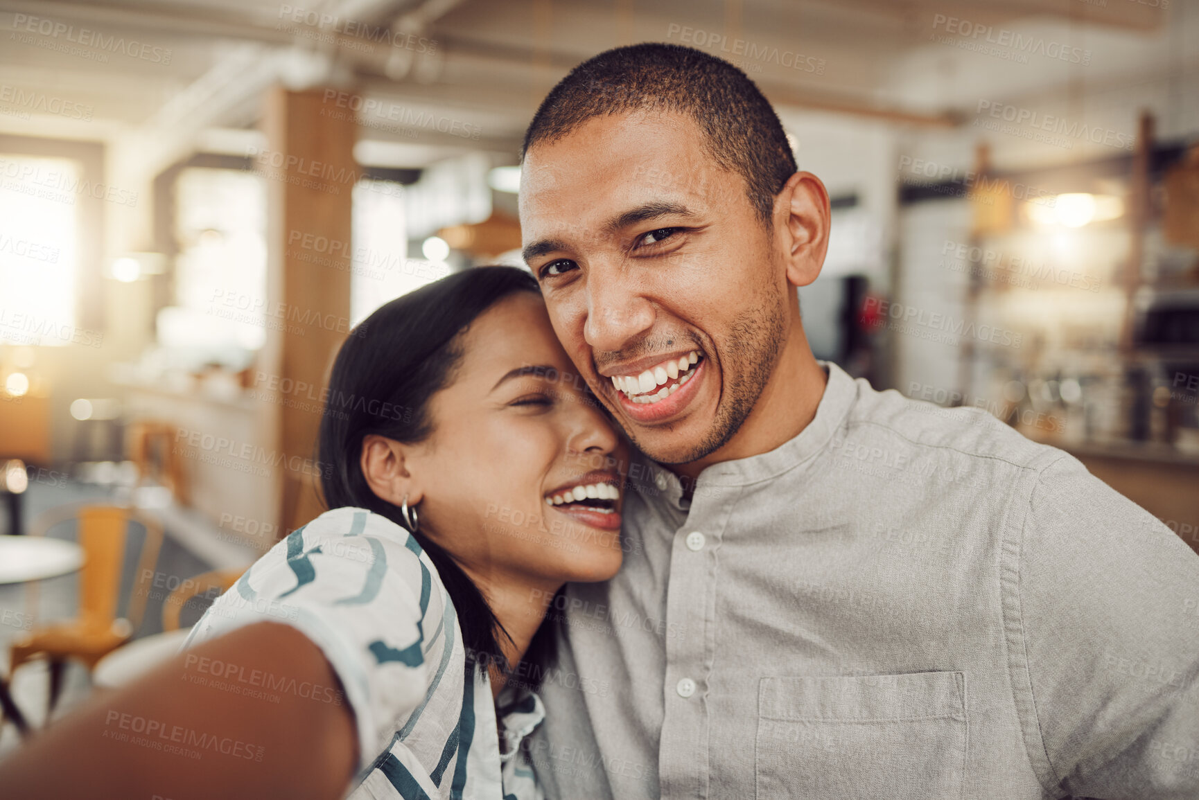 Buy stock photo Loving young laughing and having fun while holding mobile phone to take a selfie in a cafe. Happy mixed race man and woman looking happy while sitting together on a date