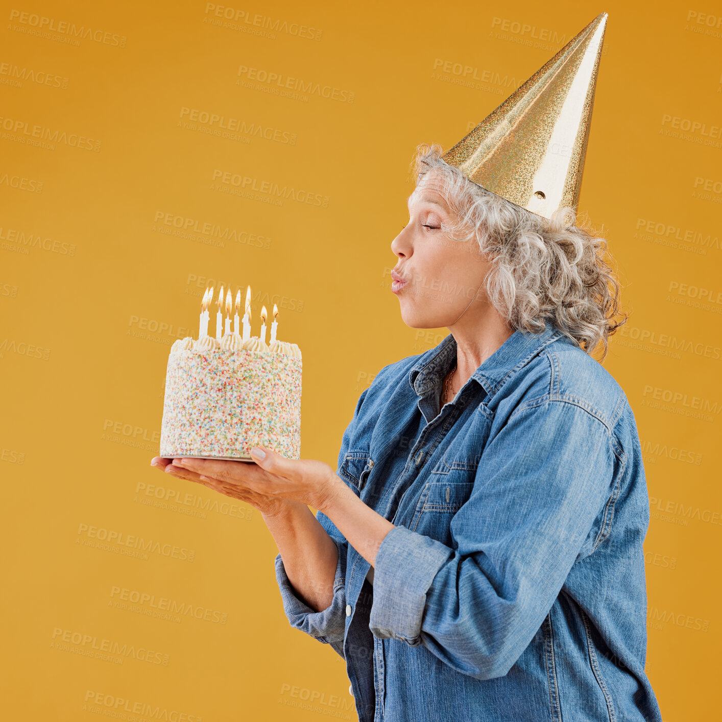 Buy stock photo One happy mature caucasian woman blowing out candles on a cake she is holding while wearing a birthday hat against a yellow background in the studio. Smiling white lady celebrating and making a wish