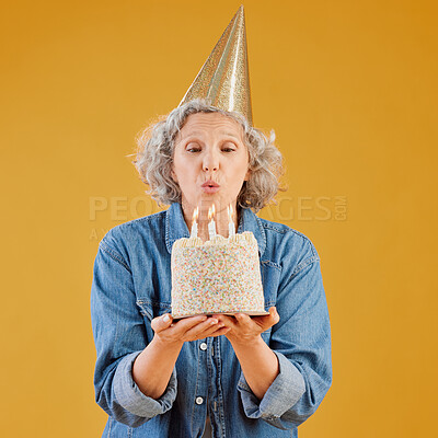 One happy mature caucasian woman blowing out candles on a cake she is holding while wearing a birthday hat against a yellow background in the studio. Smiling white lady celebrating and making a wish