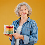 One happy mature caucasian woman holding a colourful cake with a slice missing against a yellow background in the studio. Smiling white lady showing joy and happiness while celebrating her birthday