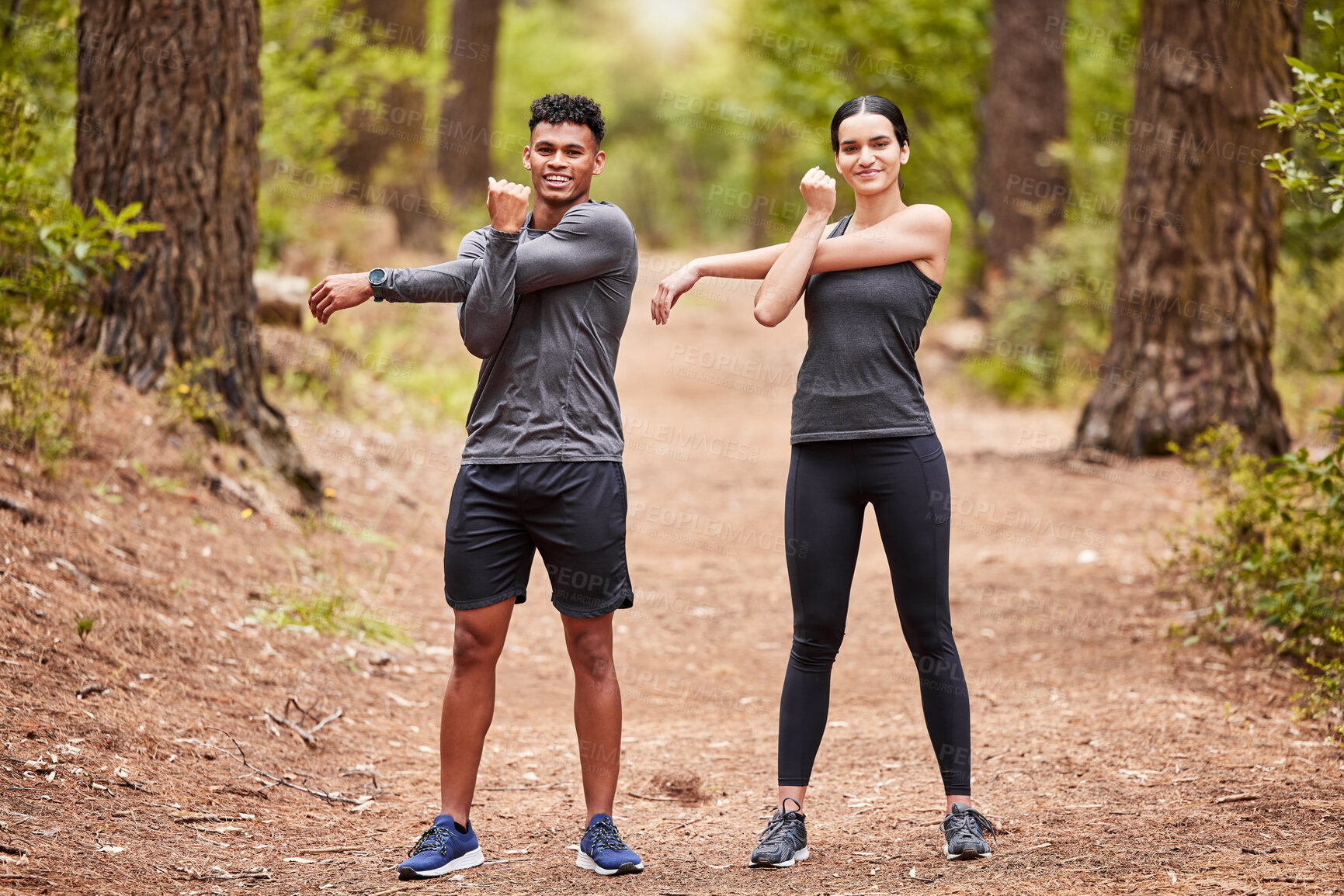 Buy stock photo Portrait of a happy young male and female athlete stretching their arms before a run outside in nature. Two fit sportspeople doing warm-up exercises in pine forest on a sunny day