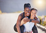 Two fit playful women wearing sportswear while out for a run outdoors. Cheerful young sportswoman giving her friend a piggyback ride on her back while exercising on a road