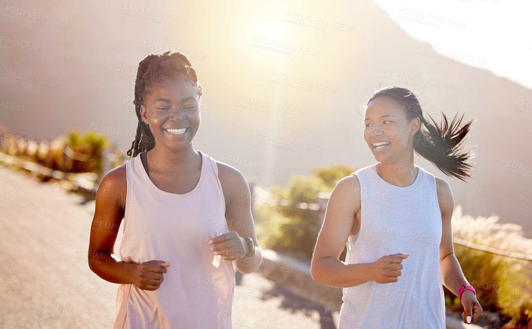 Buy stock photo Two happy young female athletes out for a run on a mountain road on a sunny day. Energetic young women running outdoors to help their bodies in shape and fit. Two diverse female friends exercising together