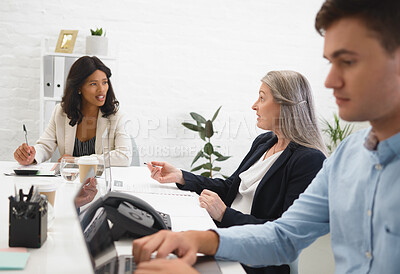 Mature caucasian businesswoman talking to an african american colleague while a young male coworker works on his laptop in an office at work. Businesswomen having a meeting at a table