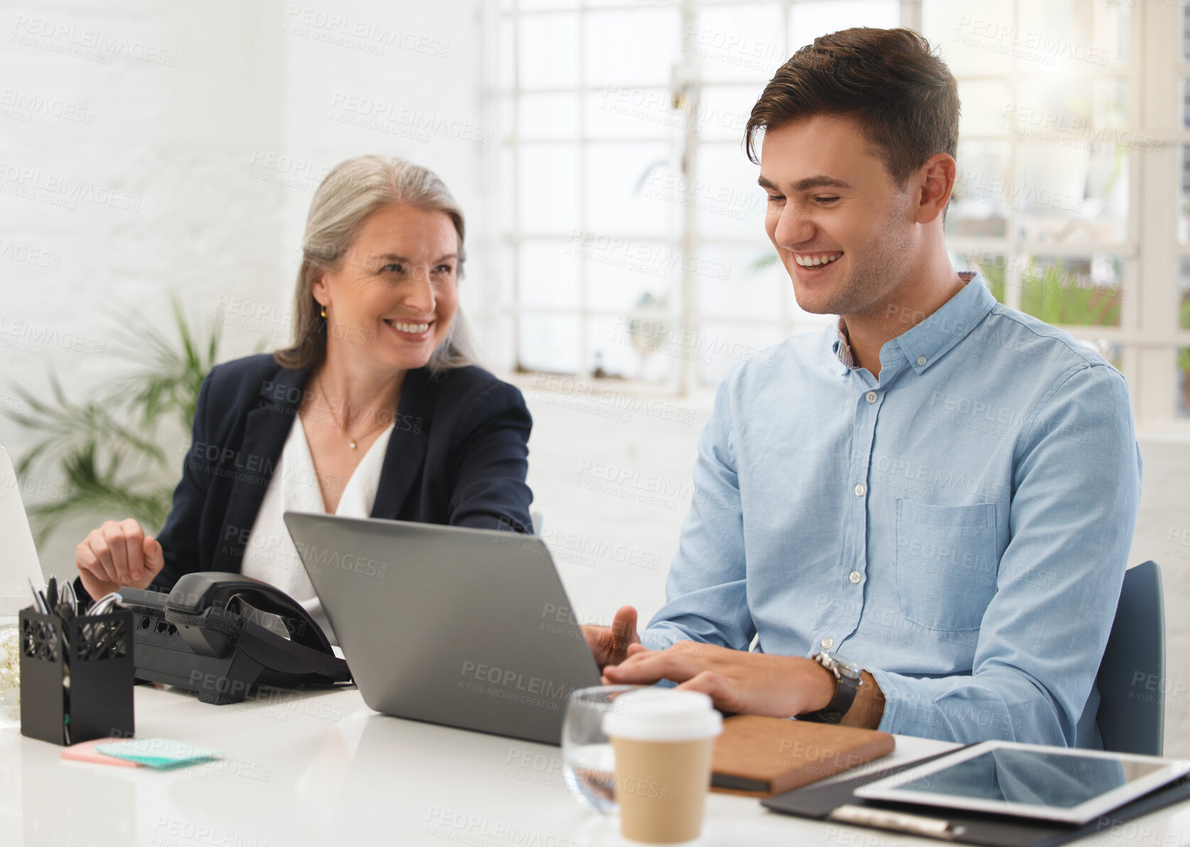 Buy stock photo Mature caucasian businesswoman talking to a young male colleague while working on a laptop together at work. Two colleagues laughing while sitting at a desk together. Businessman typing on a laptop and talking to a coworker