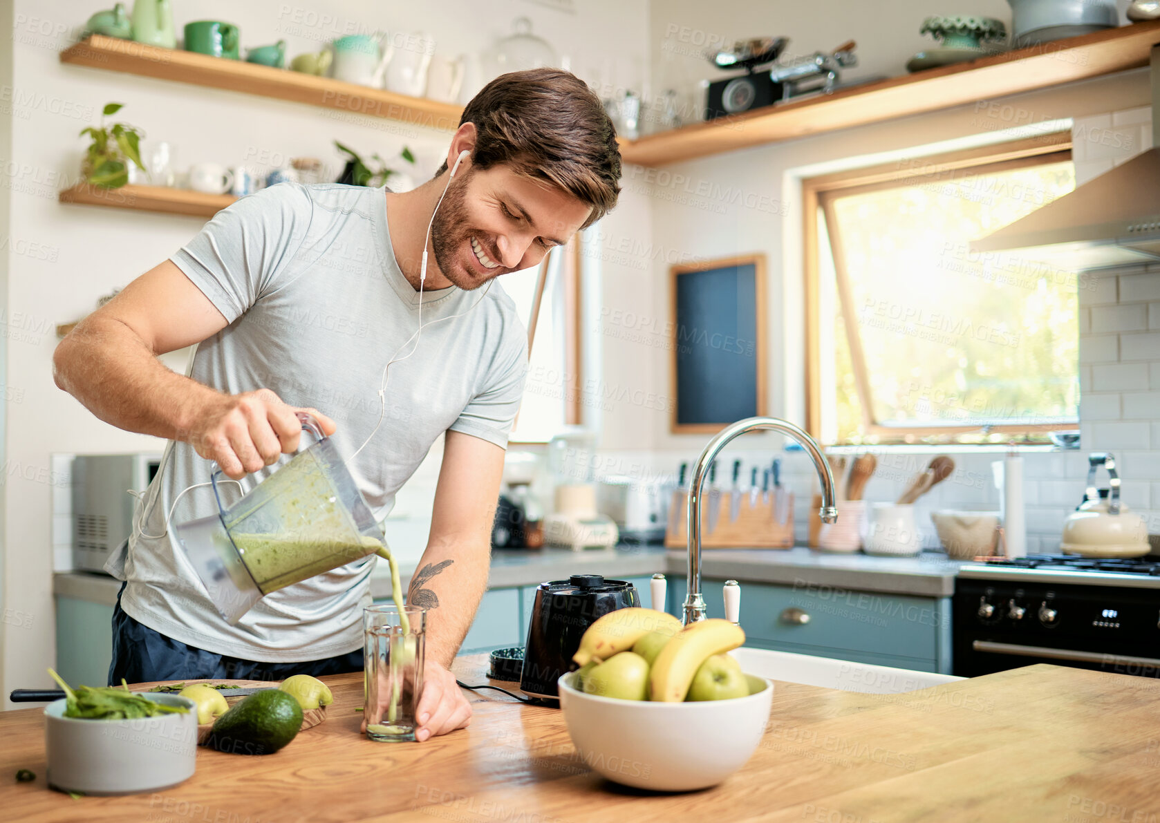 Buy stock photo One fit young caucasian man pouring healthy green detox smoothie from blender while wearing earphones in kitchen at home. Guy having fresh fruit juice to cleanse and provide energy for training. Wholesome drink with vitamins and nutrients