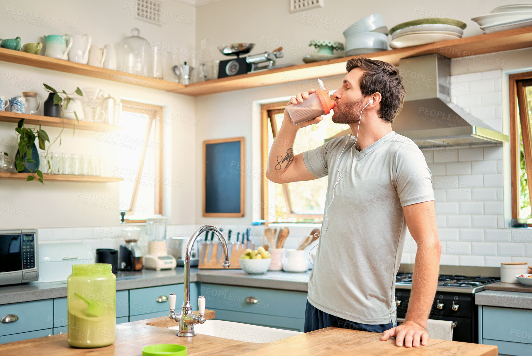 Buy stock photo One fit young caucasian man drinking bottle of chocolate whey protein shake for energy for training workout while wearing earphones in a kitchen at home. Guy having nutritional sports supplement for muscle gain and dieting with weightloss meal replacement