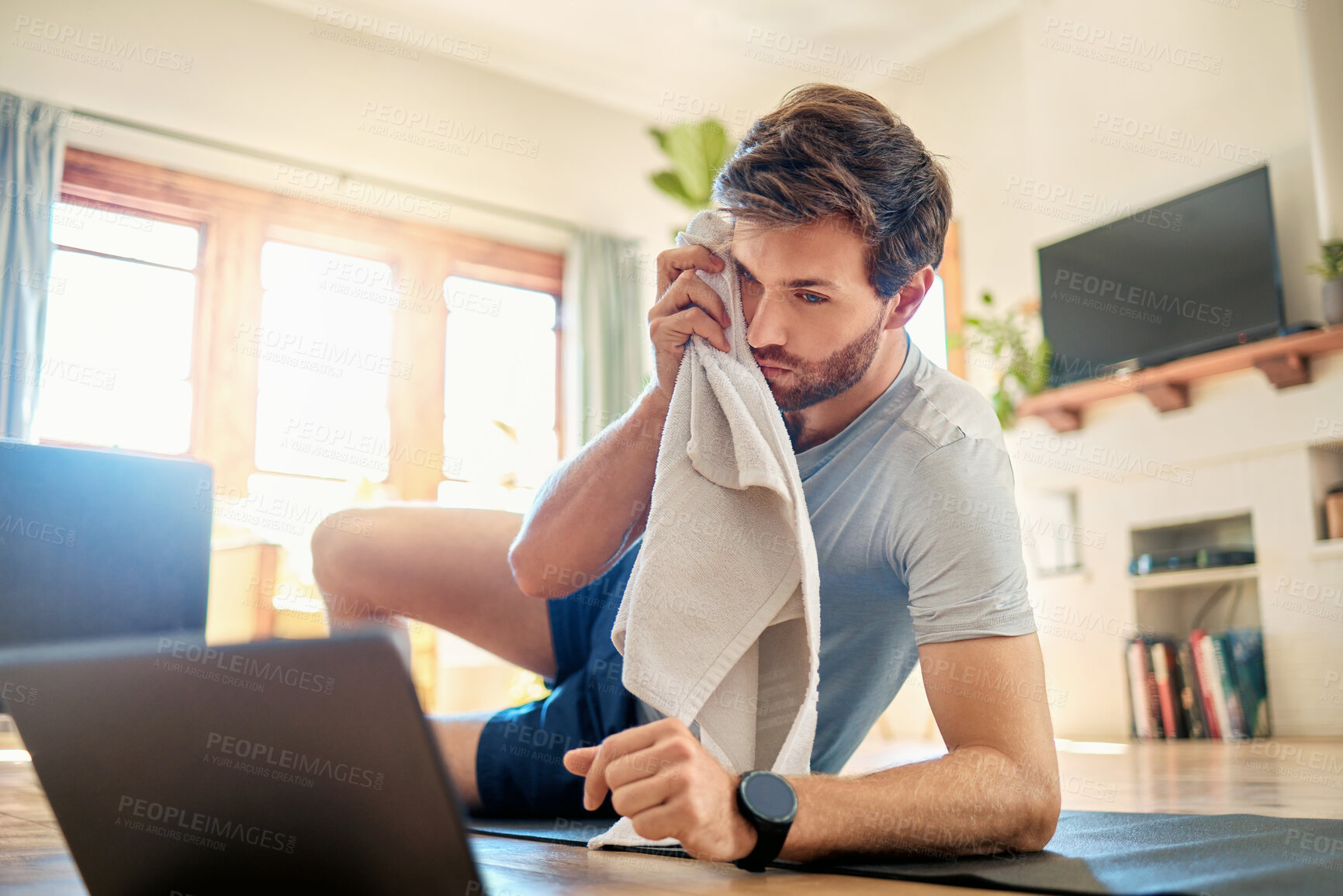 Buy stock photo One fit young caucasian man wiping sweat off forehead with a towel while taking a break from exercise at home. Guy looking tired while training with online workout tutorials on digital tablet