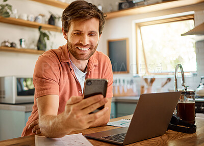 Young cheerful caucasian businessman using a phone while working on a laptop at home alone. Happy male businessperson smiling and using social media on a cellphone while working in the kitchen at home
