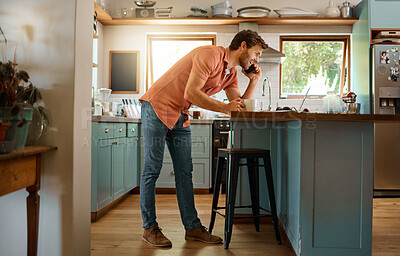 Fullbody of a young caucasian businessman on a call using a cellphone while working on a laptop and drinking coffee at home alone. Happy male businessperson talking on a cellphone while working in the kitchen at home