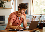 Young caucasian businessman writing in a notebook and working on a laptop at home alone. Focused male businessperson taking notes in a diary while working in the kitchen at home