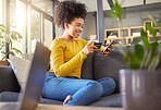 Young happy mixed race woman using a credit card and phone alone at home. Cheerful hispanic female with a curly afro making an online purchase with a debit card and cellphone while sitting on the couch at home