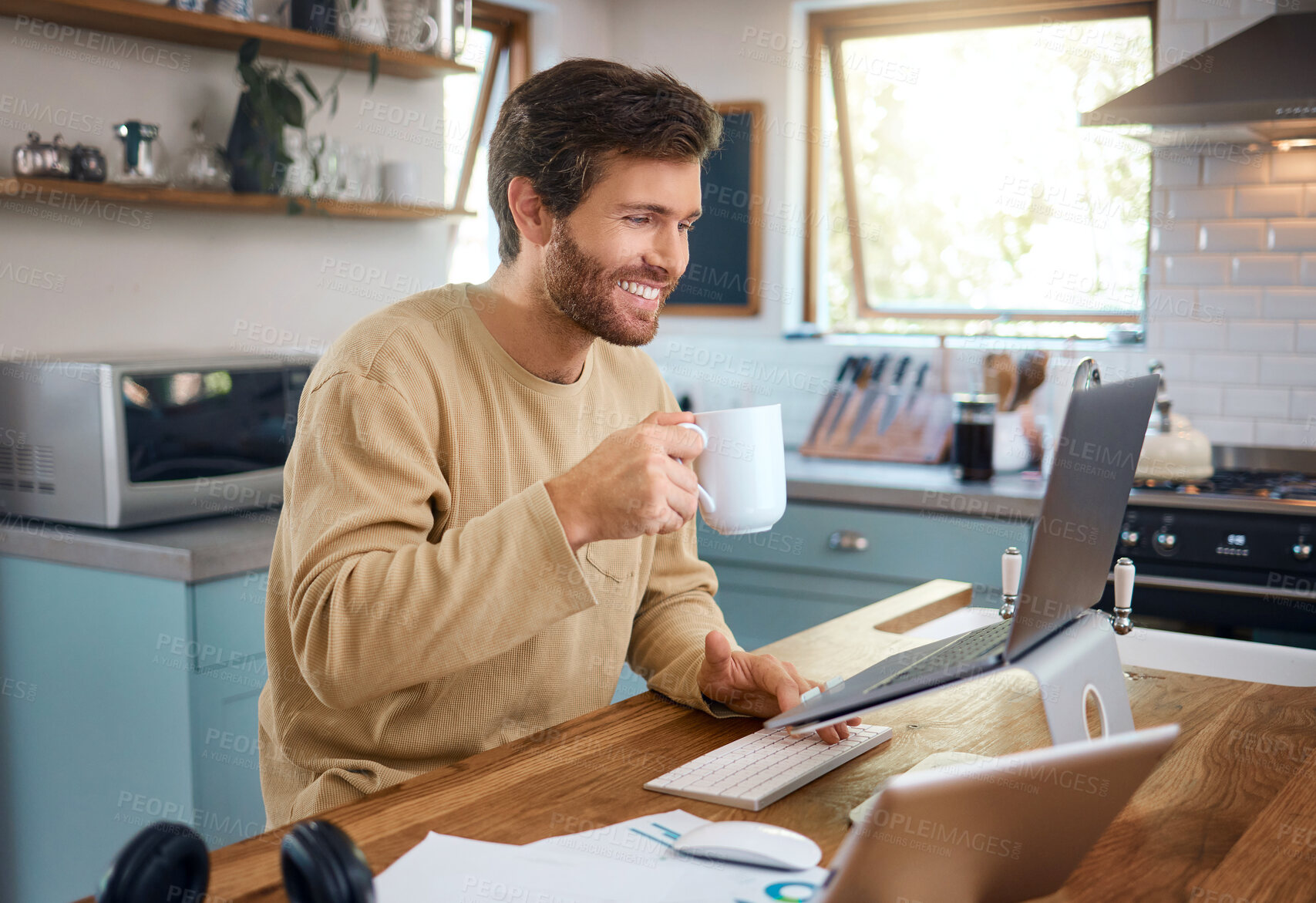 Buy stock photo Happy young caucasian man drinking coffee while working on laptop in kitchen, checking his email or searching information while doing freelance work at home. Smiling young male using internet banking service