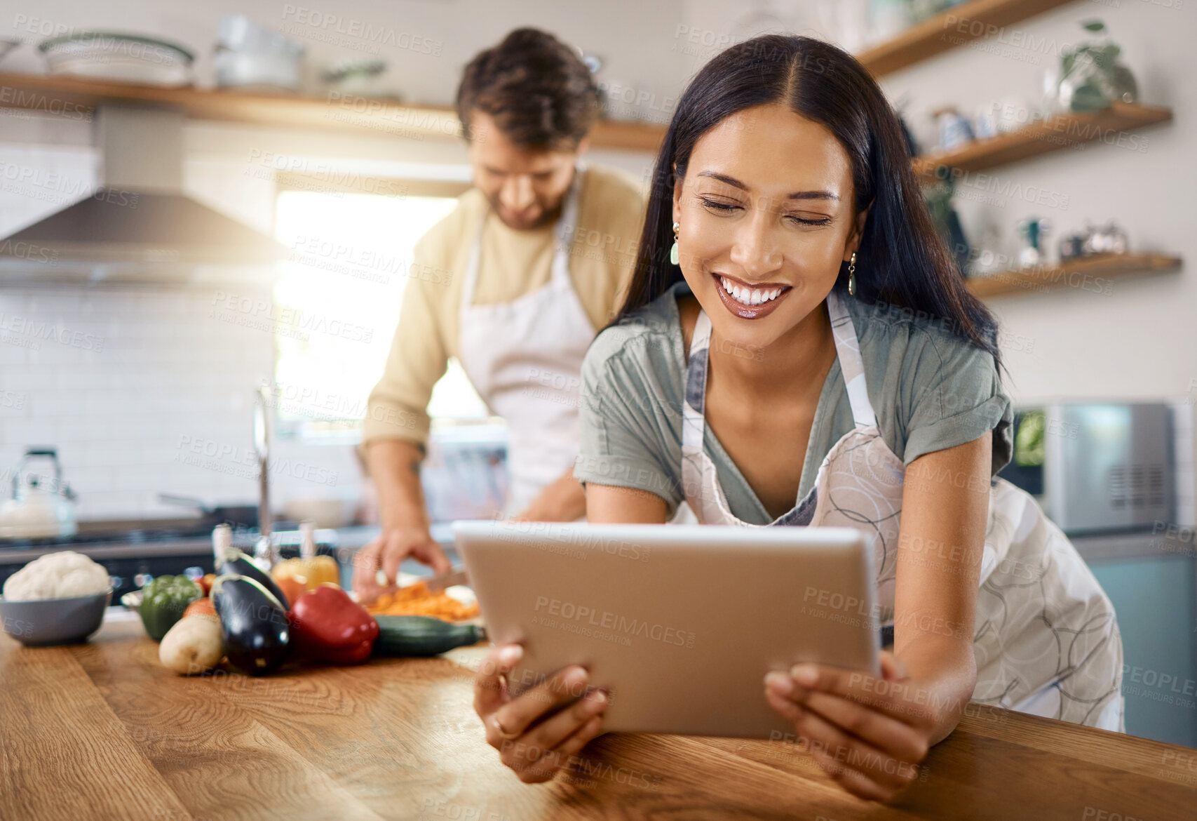 Buy stock photo Close up of happy young woman browsing on digital tablet while her boyfriend cooks in the background. Young hispanic female searching for recipe to follow online while cooking at home with her husband