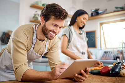Close up of happy young man browsing on digital tablet searching for recipe to follow online while cooking at home with his wife