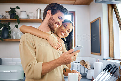 Happy young interracial couple being loving and affectionate at home. Young man using his smartphone and holds coffee cup while his girlfriend embraces him from behind. Browsing social media or sending text message