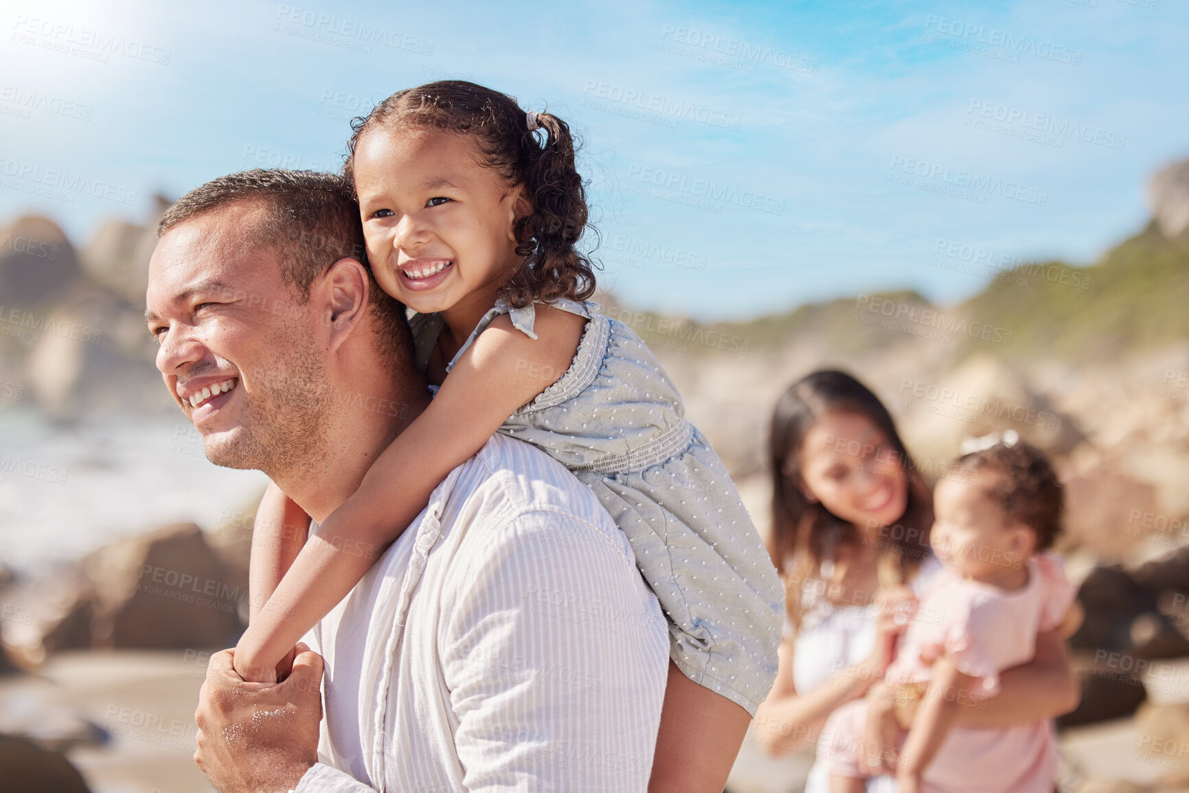 Buy stock photo A happy mixed race family of four enjoying fresh air at the beach. Hispanic couple bonding with their daughters while walking and carrying their daughters