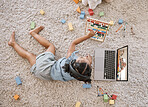 Overhead view of a little cute girl using a laptop and wireless headphones while laying on the floor in the lounge. Hispanic girl using a wireless device to do her homework in the living room