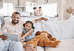 Mixed race father and daughter relaxing on the sofa in the living room while using a digital tablet while his wife looks after their other daughter in the kitchen