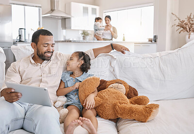 Buy stock photo Mixed race father and daughter relaxing on the sofa in the living room while using a digital tablet while his wife looks after their other daughter in the kitchen