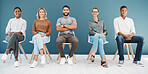 Portrait of a diverse group of confident business men and women sitting on chairs in waiting room against a blue background. Happy hopeful candidates and applicants ready for shortlist interview in line for job opportunity and vacancy