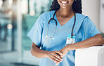 Closeup african american woman nurse smiling and leaning against a wall while standing in hospital. She's here to take care of her patients' wellbeing. Health and safety in the field of medicine