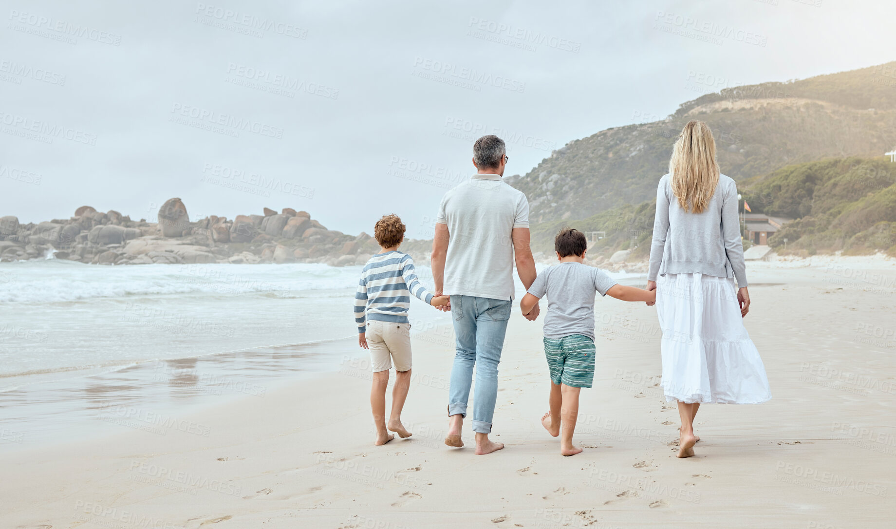 Buy stock photo Happy caucasian parents and kids from the back holding hands while walking together along the beach shore during a relaxing fun family summer vacation. Loving mom and dad enjoying leisurely stroll while bonding with little sons