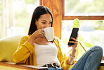 Woman using her smartphone and drinking coffee sitting in front of a window in a bright living room. A young female looking at her cellphone relaxing on a chair using modern technology at home