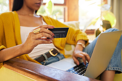 Unrecognizable woman shopping online while typing on her laptop and holding her bank card sitting on a chair in a bright living room. A hispanic young female at home using modern technology for buying