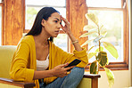 Worried woman at home sitting on a chair in a bright living room with her hand against her head holding a smartphone. One tired and anxious young female inside with a headache and stress